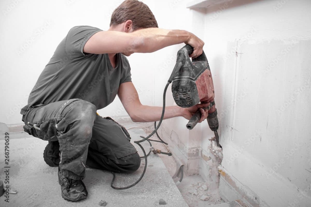 Ein Arbeiter stemmt eine Wand mit dem Bohrhammer auf. In Eigenleistung schafft er so die Öffnung für eine Heizungsleitung.