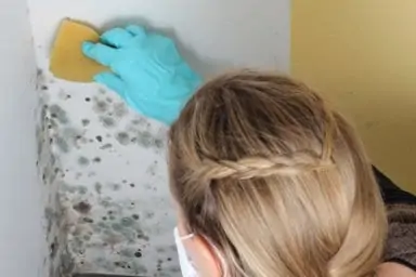 Frau entfernt Schimmel in der Ecke mit Mundschutz und Gummihandschuhen.